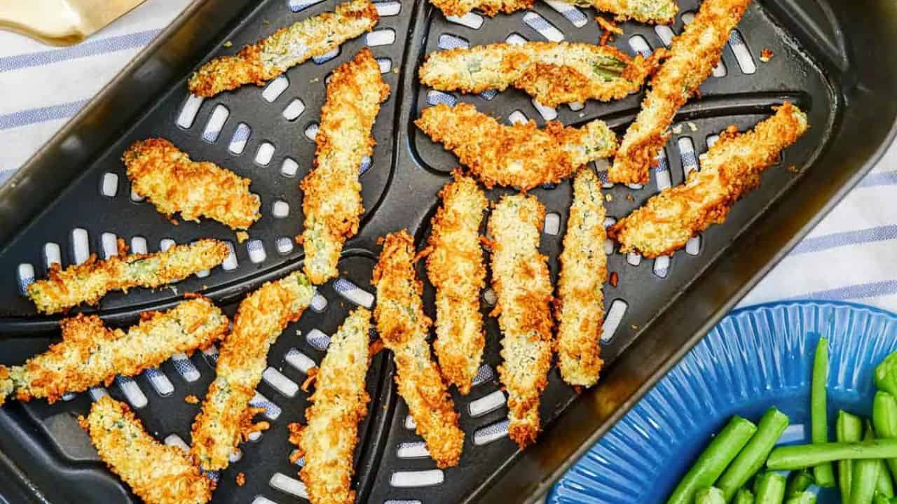 Breaded zucchini fries in an air fryer basket, golden and crispy, with a blue plate of green beans visible beside the air fryer.