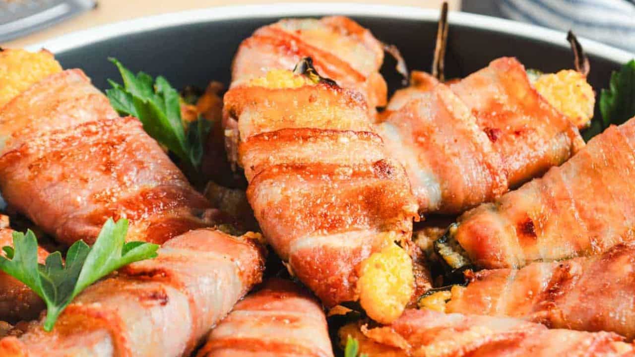Plate of bacon-wrapped peppers filled with cheese, garnished with parsley, next to a grated cheese block and grater on a wooden board.