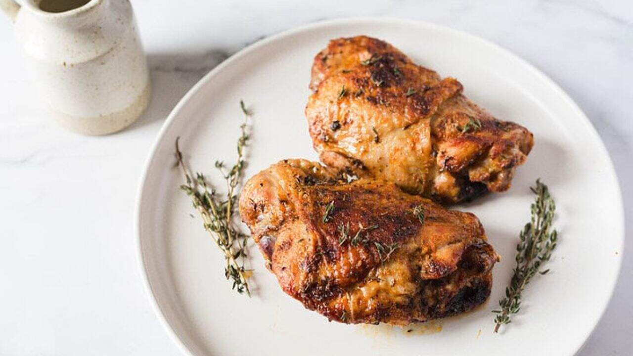 Two roasted chicken thighs on a white plate garnished with sprigs of thyme. A ceramic jar is partially visible in the background on the left.