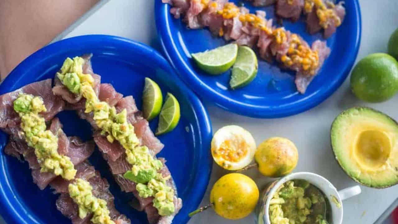 Slices of raw fish topped with diced avocado are arranged on a blue plate, with slices of lime and whole passion fruits visible in the background.