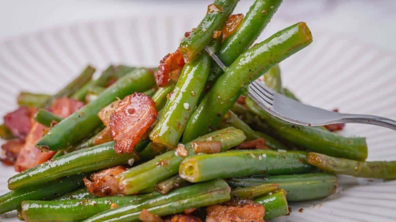 Close-up of a plate with sautéed green beans and bacon pieces, with a fork spearing a few beans.