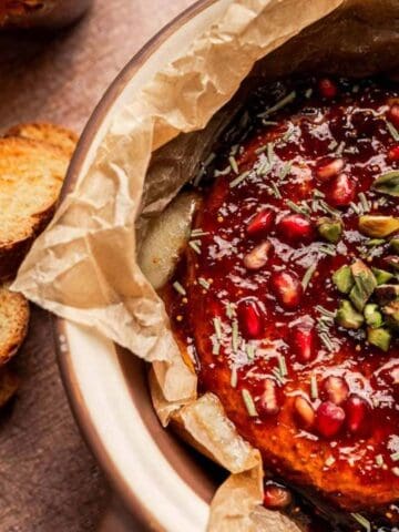 Round baked cheese topped with pomegranate seeds, chopped pistachios, and herbs, served on parchment paper with crostini on the side.