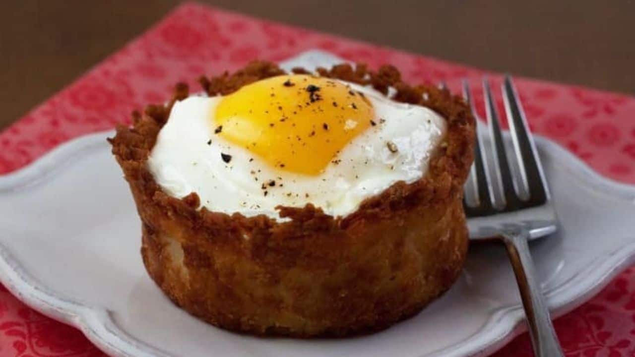 A breakfast dish: crispy potato nest with a sunny-side-up egg on top, seasoned with black pepper. A fork is placed on the right side of the white plate.