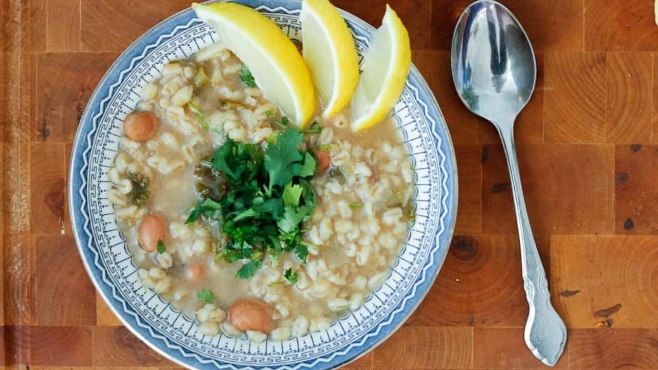 A bowl of barley soup with lemon wedges on a wooden table.