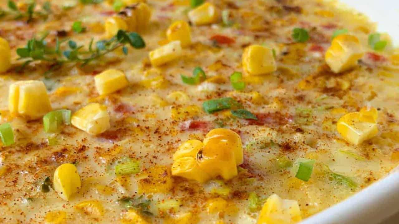 A baked corn casserole with chopped peppers and herbs is presented in a white oval dish on a wooden surface, with a patterned cloth nearby.