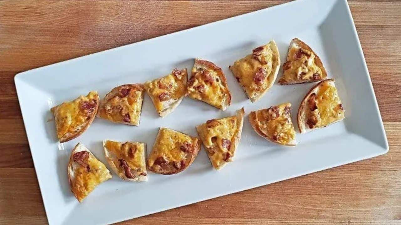 Image shows a hand holding a quarter of a Boozy Bacon Cheddar Bite with more on a white tray in the background.