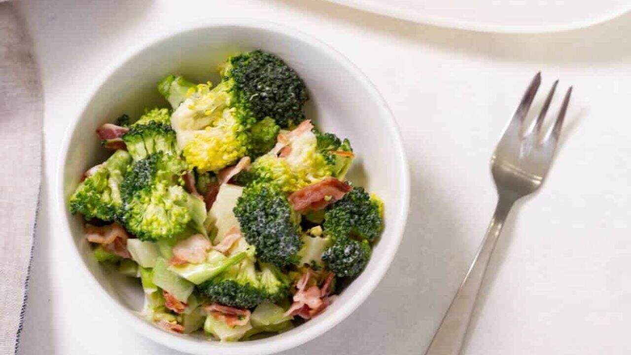 Broccoli salad with bacon bits on a white oval plate, accompanied by a wooden serving spoon on a linen napkin.