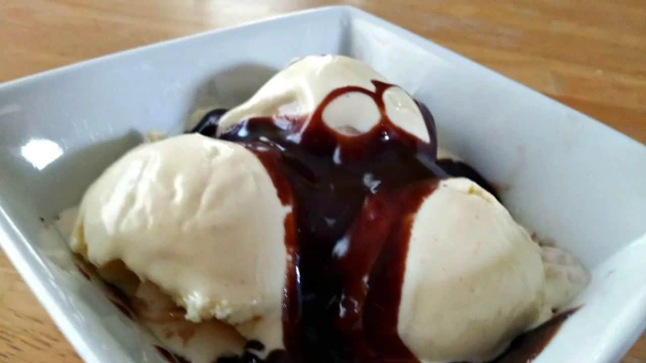 Image shows A bowl of vanilla ice cream topped with chocolate syrup on a wooden table.