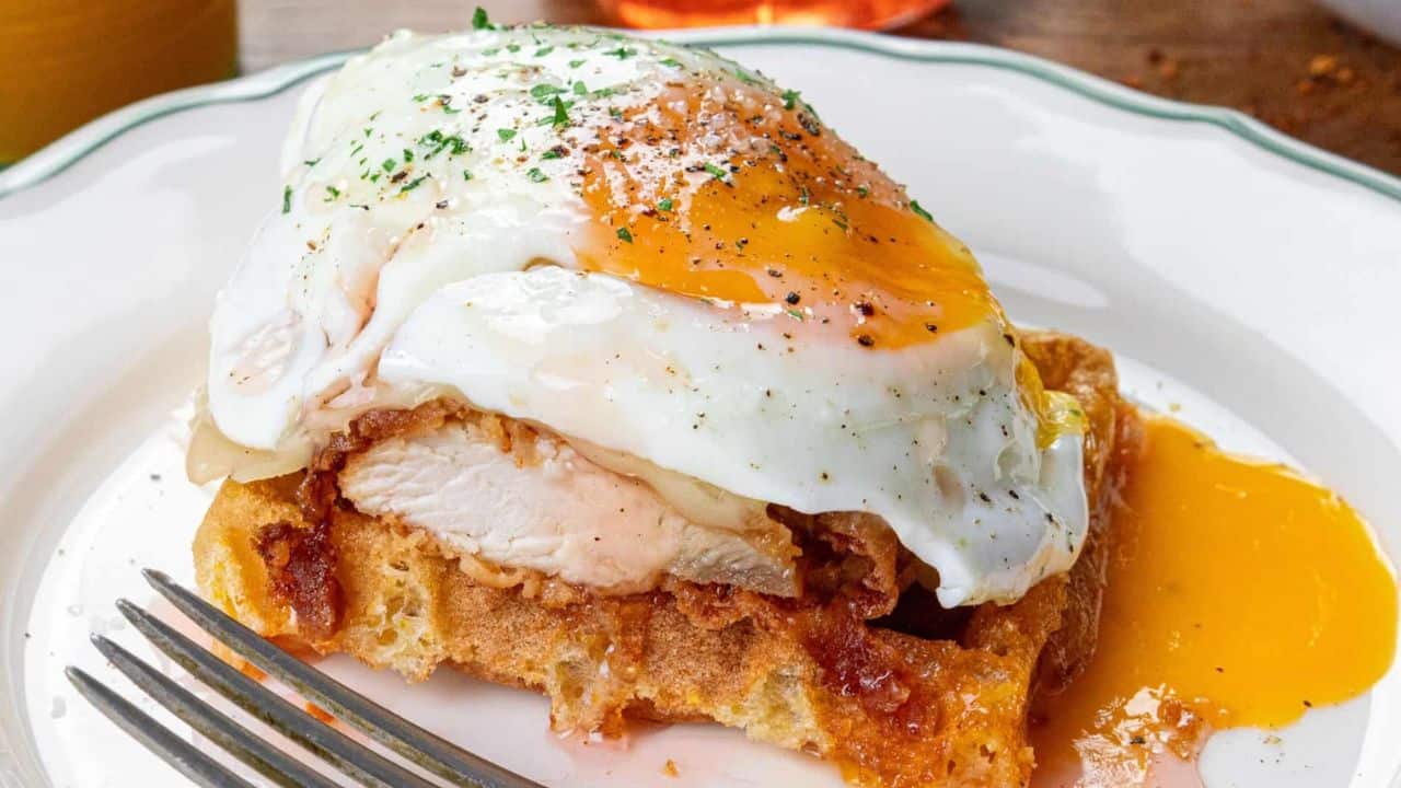 A close-up of a plate with a crispy waffle topped with fried chicken, melted cheese, and a sunny-side-up egg garnished with herbs. A fork rests on the plate's edge. In the background, there are blurred images of drinks and a small bowl.