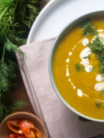 A bowl of creamy soup garnished with fresh dill, sliced red peppers, and a drizzle of cream sits on a cloth next to fresh dill leaves.