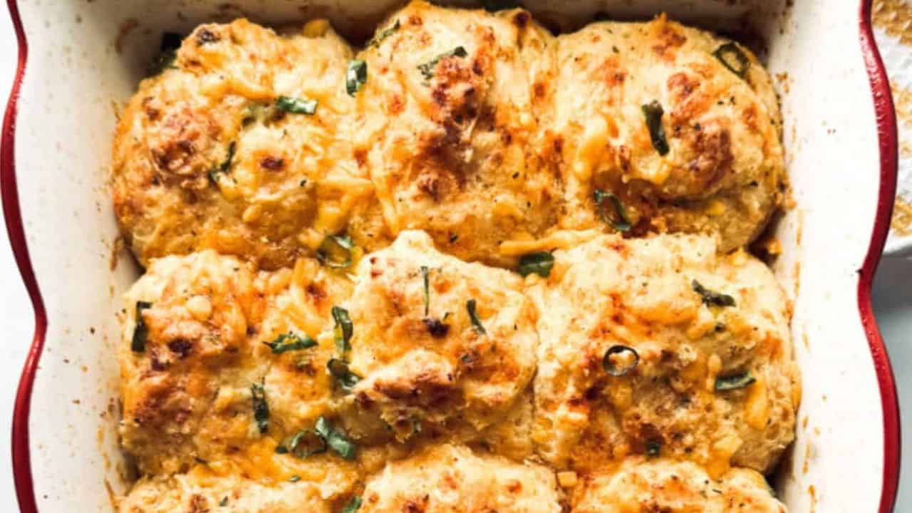 Golden-brown cheesy biscuits, sausage, and eggs topped with chopped herbs in a red floral-patterned baking dish.