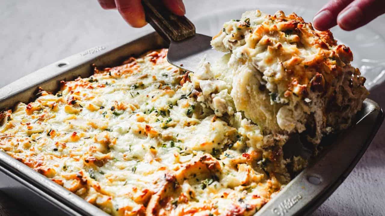 A baked casserole dish filled with a cheesy, golden-brown topping is being scooped with a spatula. The dish appears to contain layers of ingredients beneath the melted cheese, with herbs sprinkled on top for garnish.