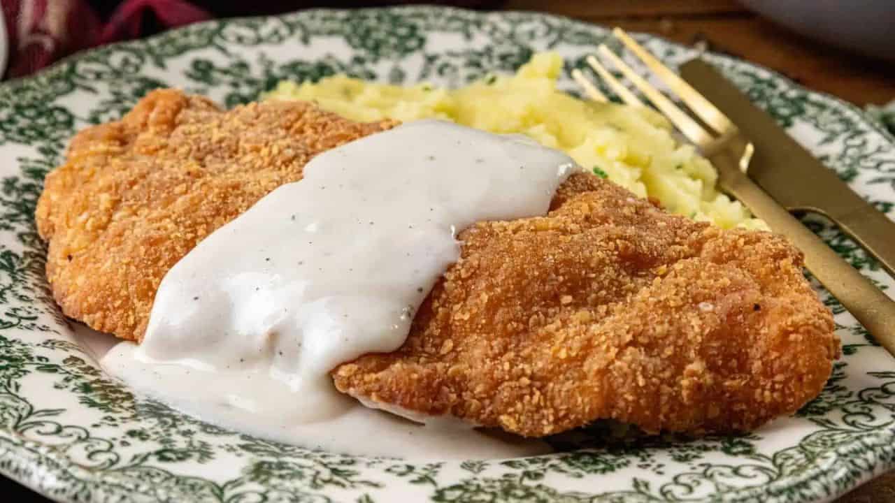 A classic Southern comfort food, this Chicken Fried Chicken features a breaded and fried cutlet topped with rich gravy, served with mashed potatoes on a decorative green and white plate beside a gold fork and knife.