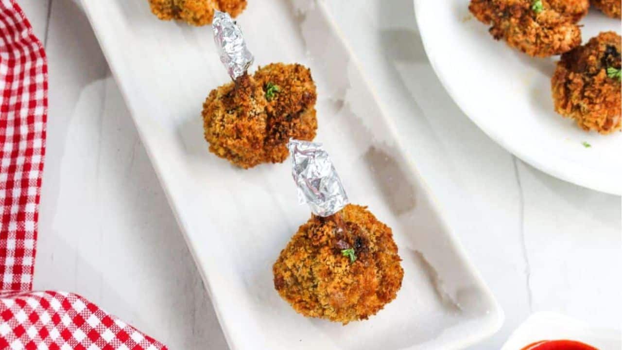 A close-up of a hand holding a crispy breaded chicken drumstick, with more drumsticks on a white plate in the background.