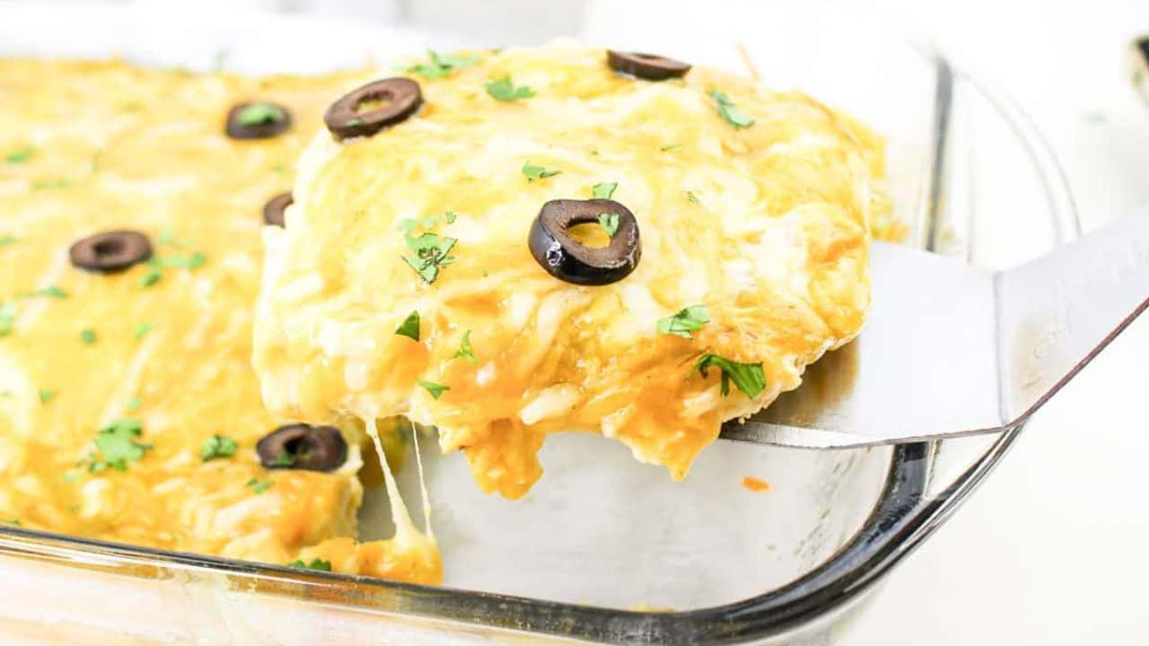 A slice of cheesy enchilada casserole with black olives and herbs is lifted from a glass baking dish.
