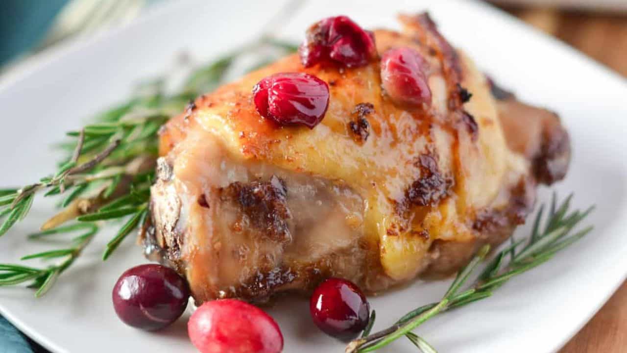 A piece of roasted chicken thighs with cranberry sauce, garnished with rosemary, rests elegantly on a white plate.