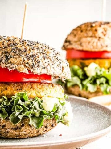 A veggie burger with lettuce, tomato, and sauce on a whole grain bun sits on a plate, with another burger and tomatoes in the background.