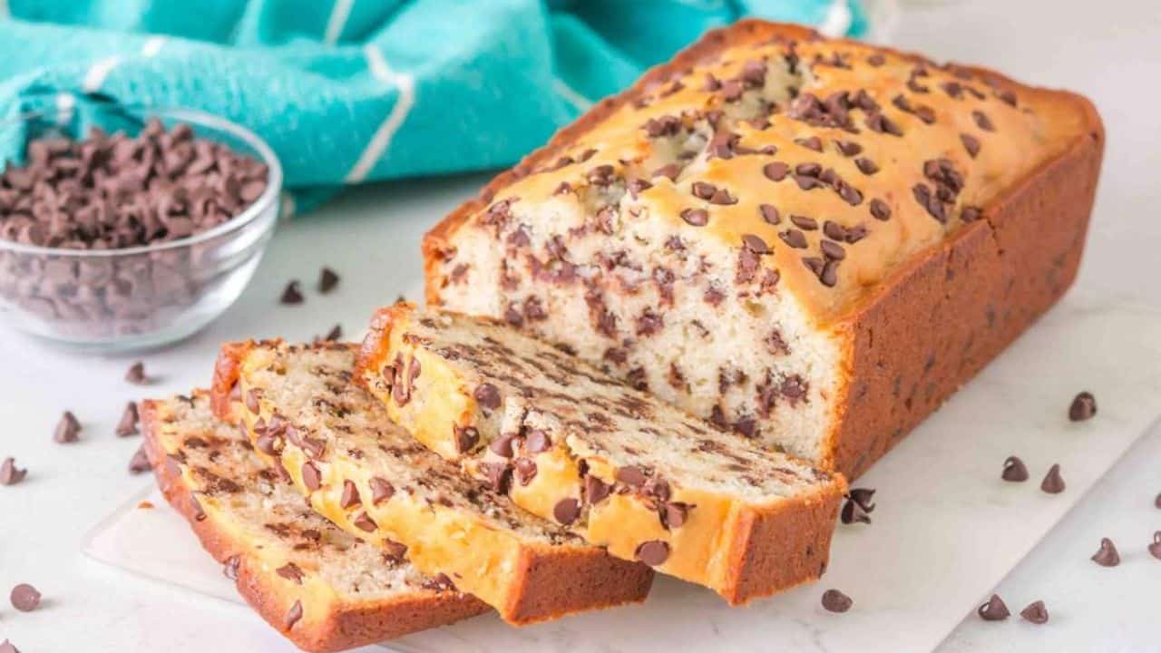 A loaf of chocolate chip bread with several slices cut, placed on a white surface. A bowl of chocolate chips and a blue patterned cloth are beside it.