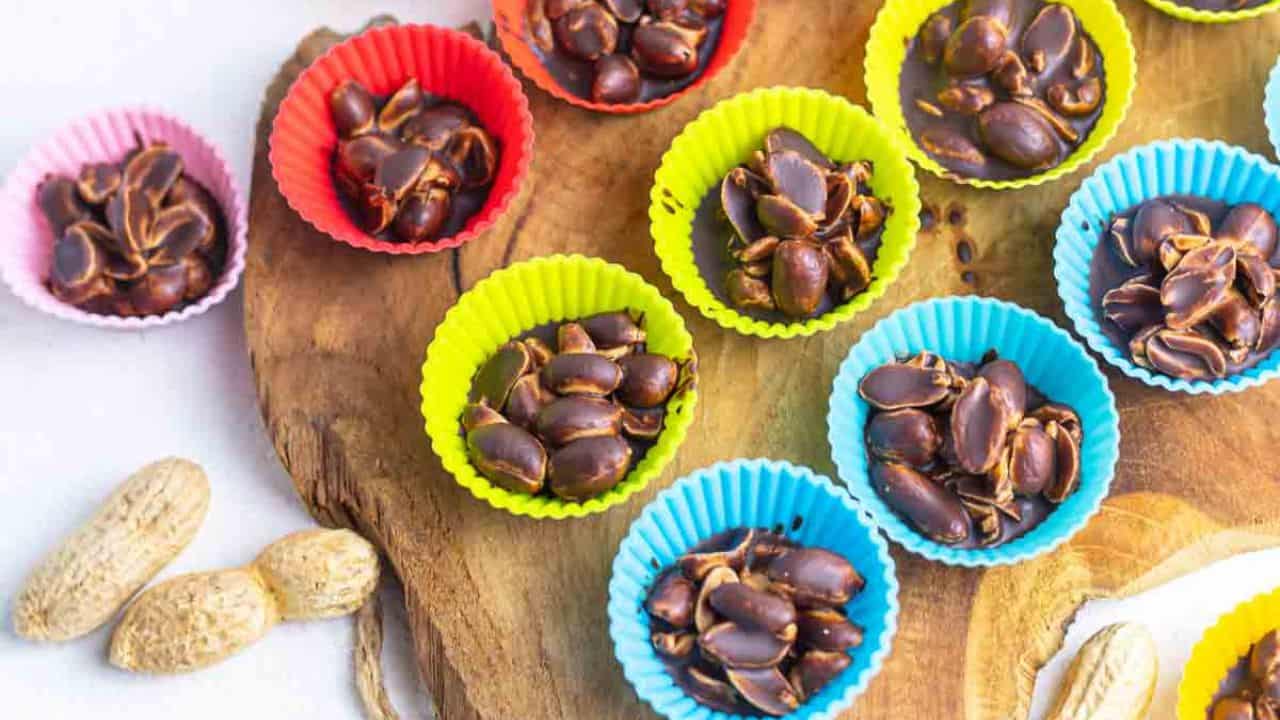 Colorful mini cupcake liners filled with chocolate and peanuts on a wooden board, surrounded by raw peanuts.