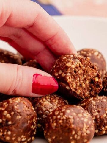 A hand with red nail polish picks up a chocolate energy ball from a white plate filled with similar balls.