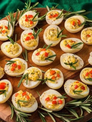 A wooden platter with deviled eggs garnished with chopped vegetables and rosemary sprigs, set on a rustic table with green cloth and garlic in the background.