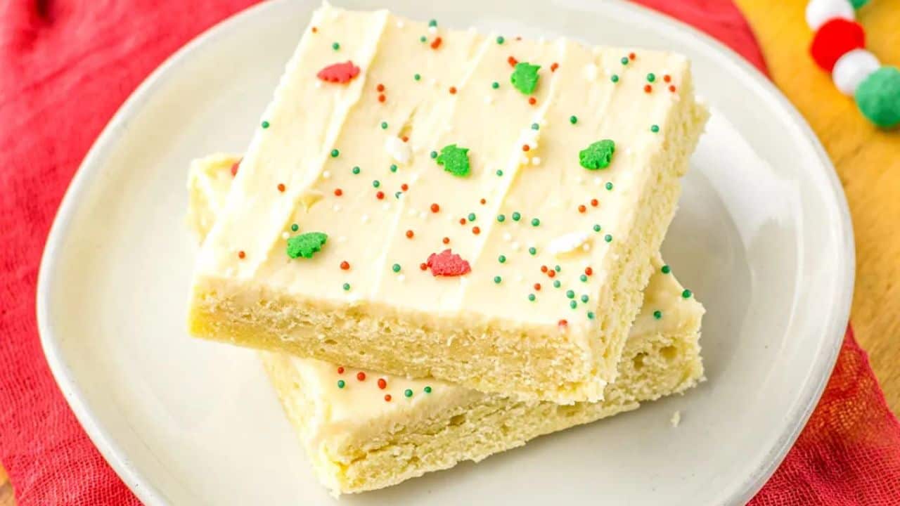 Two frosted sugar cookie bars with white icing and red, green, and white sprinkles, stacked on a white plate.