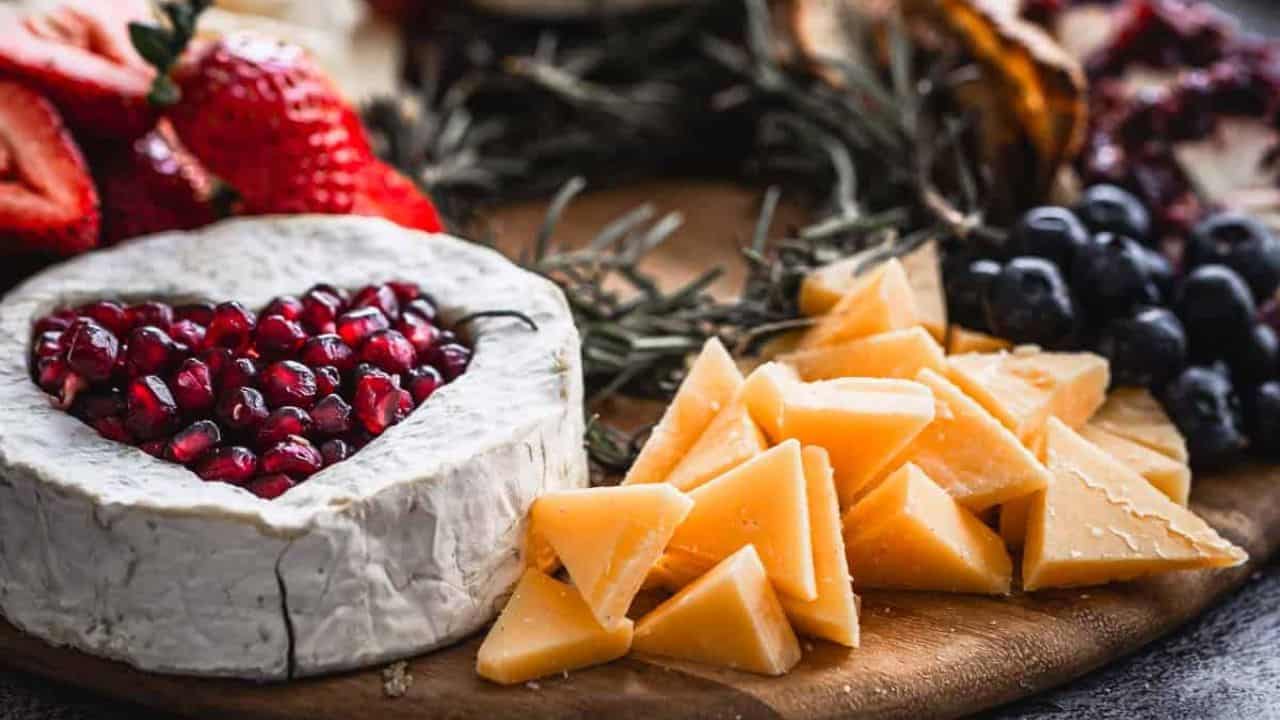A charcuterie board with heart-shaped cheese topped with pomegranate seeds, surrounded by sliced strawberries, blueberries, grapes, assorted cheeses, crackers, and pine boughs.