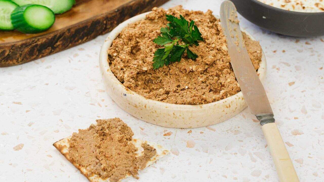 A bowl filled with a brown, chunky spread garnished with a sprig of parsley. A knife lies beside the bowl and slices of cucumber and crackers are in the background on a light-colored surface.
