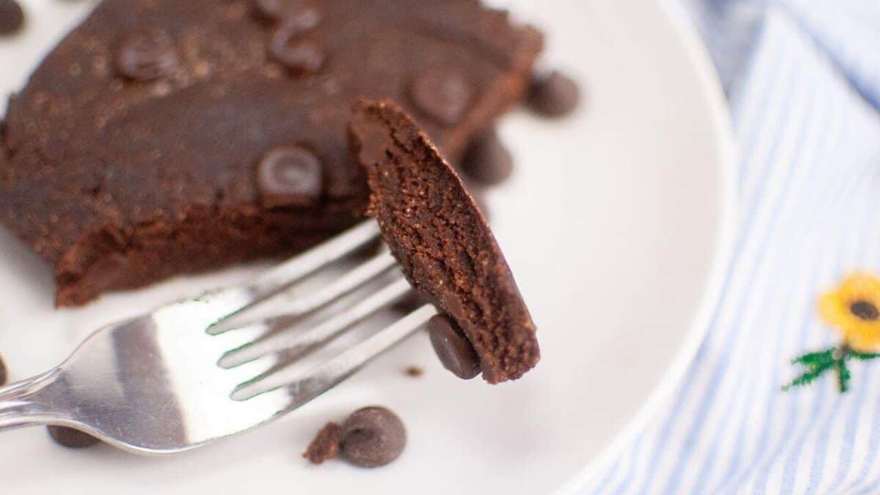 Fork holding a slice of a chocolate brownie with chocolate chips on a white plate.