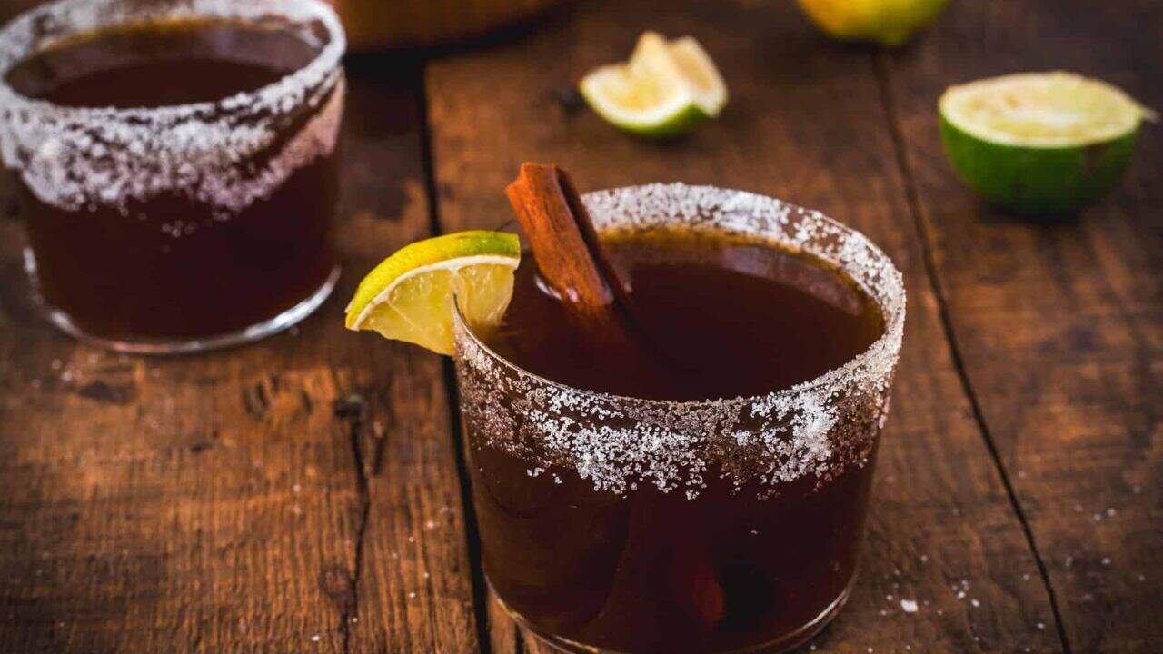 A glass filled with a dark beverage, garnished with a lime wedge and cinnamon stick, and rimmed with coarse salt. Another drink and halved limes are on a rustic wooden surface in the background.