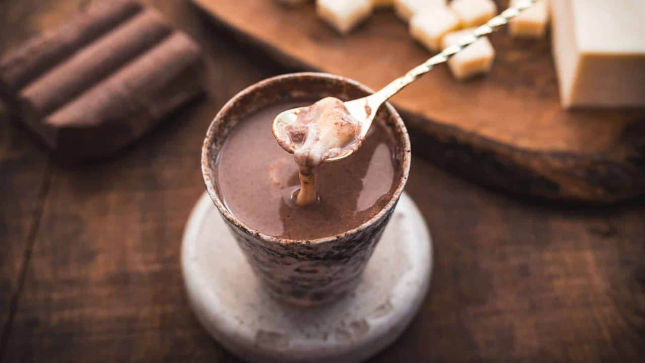 A spoon holding a thick, creamy chocolate mixture hovers over a cup filled with hot chocolate, set on a coaster on a wooden table. Chocolate blocks and a cutting board are visible in the background.