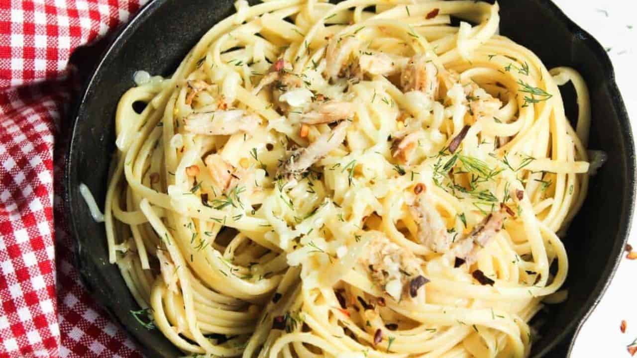 A close-up of spaghetti twirled on a fork with bits of crab meat and herbs, with more pasta and a dish in the blurred background.