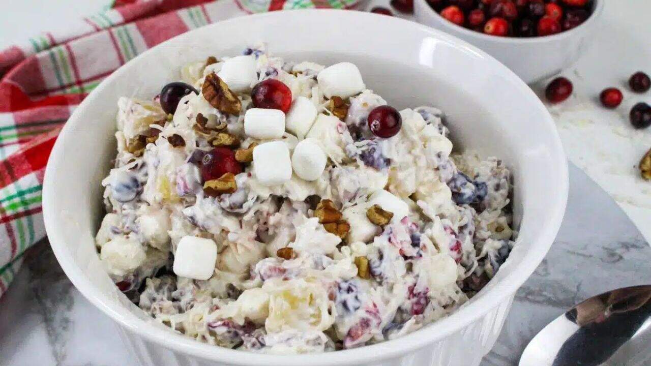 A white bowl of cranberry fluff salad topped with mini marshmallows, pecans, and cranberries on a marble surface, with a spoon and a plaid cloth nearby.