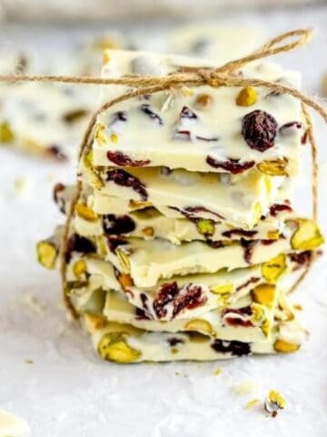 A stack of white chocolate bark pieces with pistachios and dried cranberries, tied with twine, sits on crumpled parchment paper.