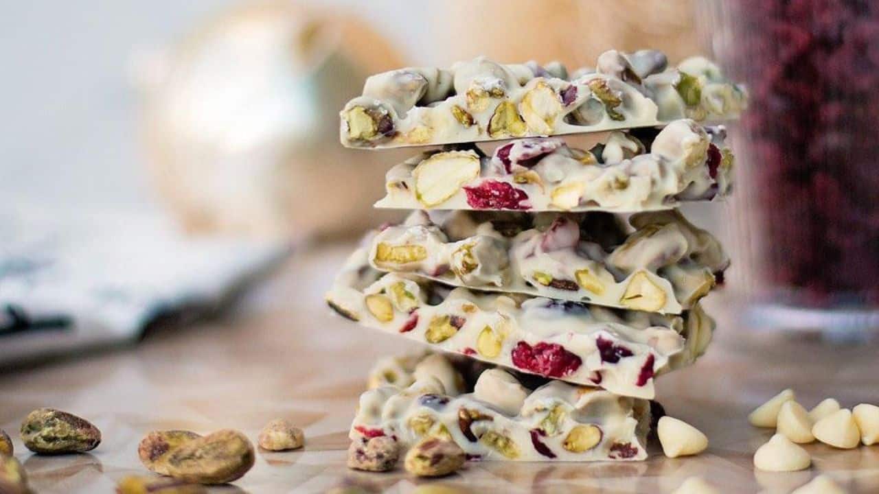 A stack of white chocolate bark pieces with pistachios and dried cranberries, placed on a wooden surface with scattered nuts and chocolate chips nearby.