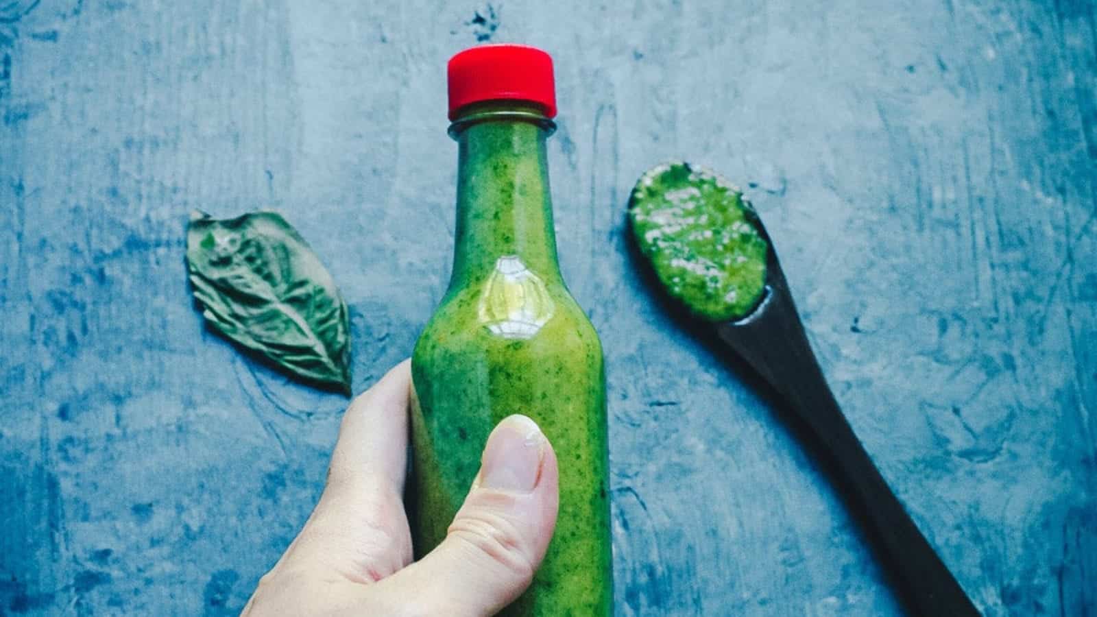 A hand holds a small glass bottle with green sauce and a red cap; a wooden spoon with green sauce and a basil leaf are nearby on a gray surface.