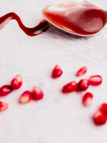 A spoon with red sauce rests on a light surface, with sauce spilled beside it and scattered pomegranate seeds in the foreground.