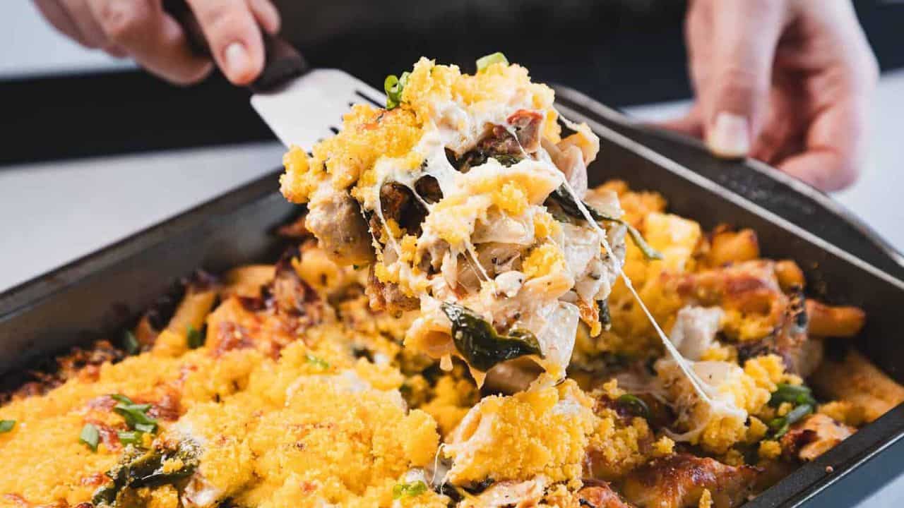 A close-up of a baked pasta dish in a rectangular metal pan with a golden breadcrumb topping, pieces of pasta, baked cheese, and green herbs visible.
