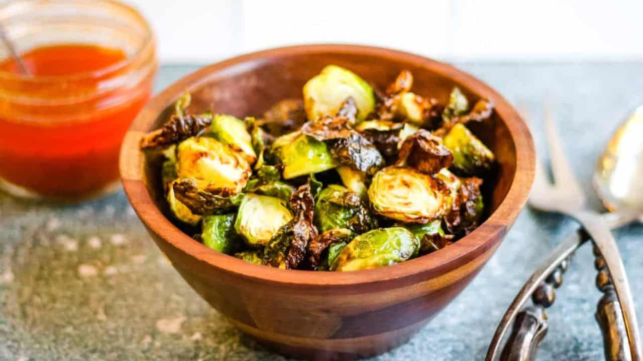 A wooden bowl filled with roasted Brussels sprouts. A jar of red sauce and a pair of tongs are in the background.