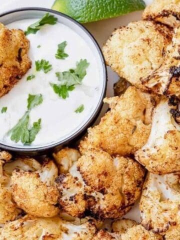 A plate of roasted cauliflower florets with a small bowl of creamy dipping sauce garnished with cilantro, and a lime wedge on the side.