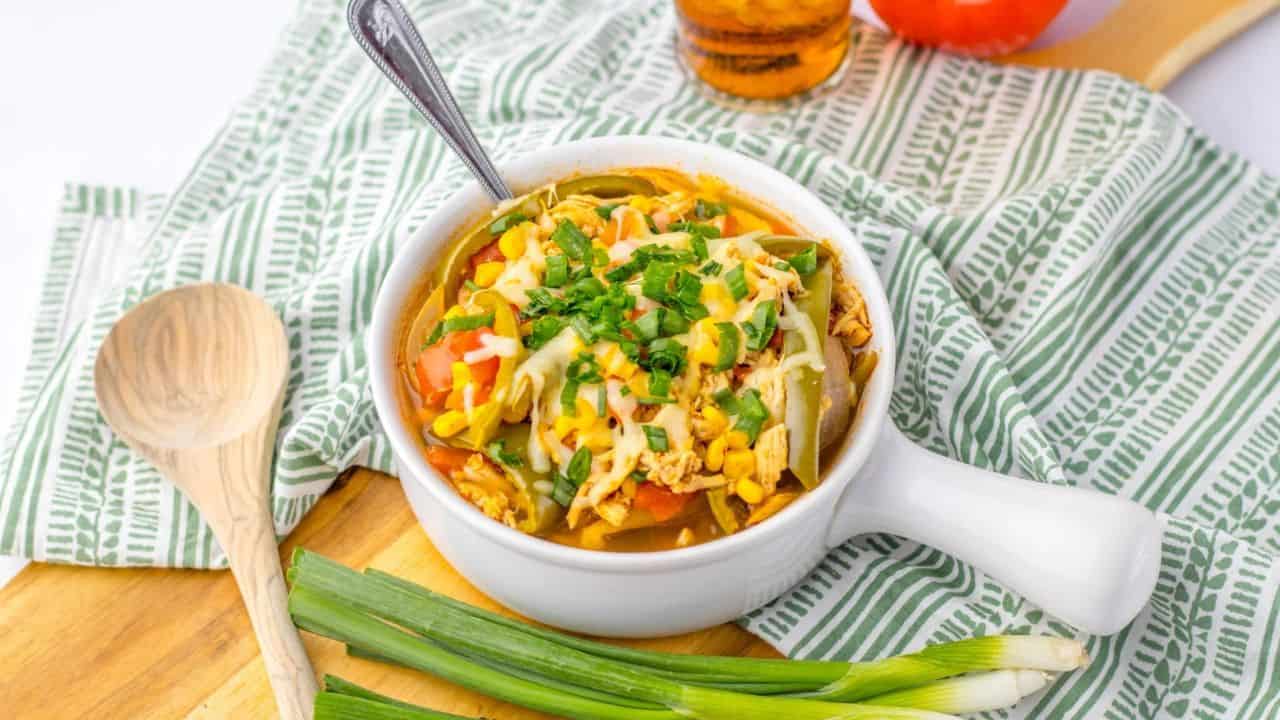 A white bowl of vegetable soup topped with chopped herbs and cheese, placed on a wooden board with green onions, a tomato, and a wooden spoon nearby.