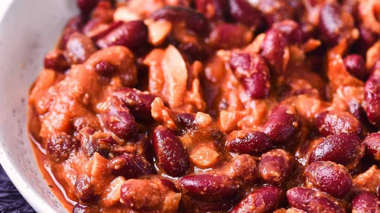 A white bowl filled with a red kidney bean stew, including visible pieces of meat or vegetables, sits on a dark cloth napkin with a fork and spoon beside it. A small bunch of parsley is on the table nearby.
