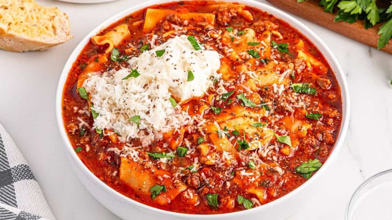 A bowl of lasagna soup topped with ricotta cheese, grated Parmesan, and parsley. Slices of bread and fresh parsley sit nearby on a white surface.