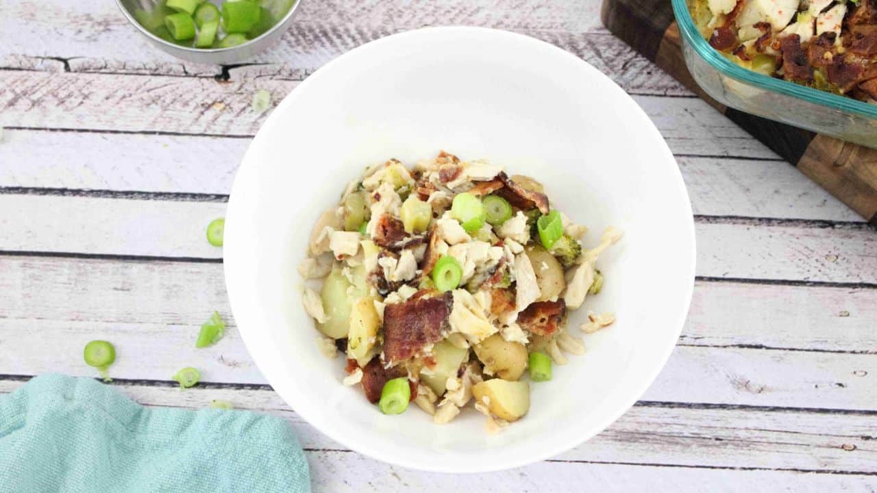 A white bowl filled with a potato salad topped with bacon pieces, shredded cheese, and sliced green onions sits on a light wooden surface.