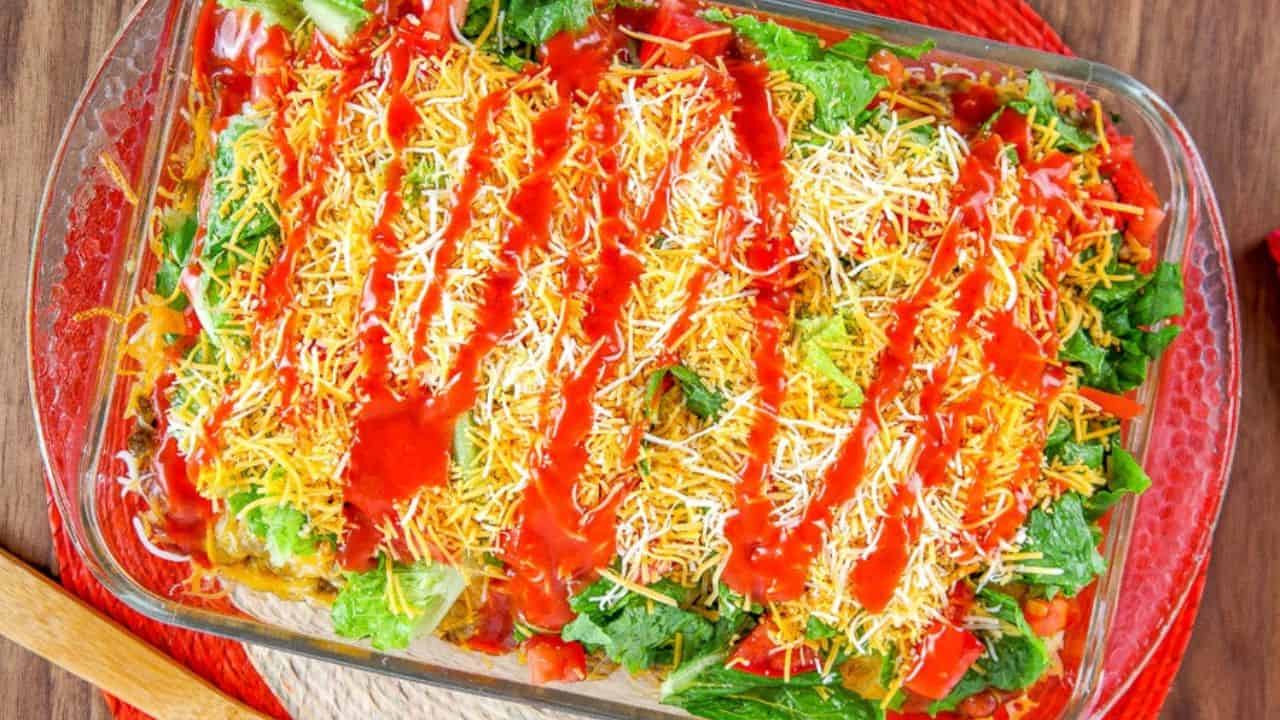 A glass baking dish filled with layered salad ingredients, topped with shredded cheese, lettuce, and drizzled with a red sauce.