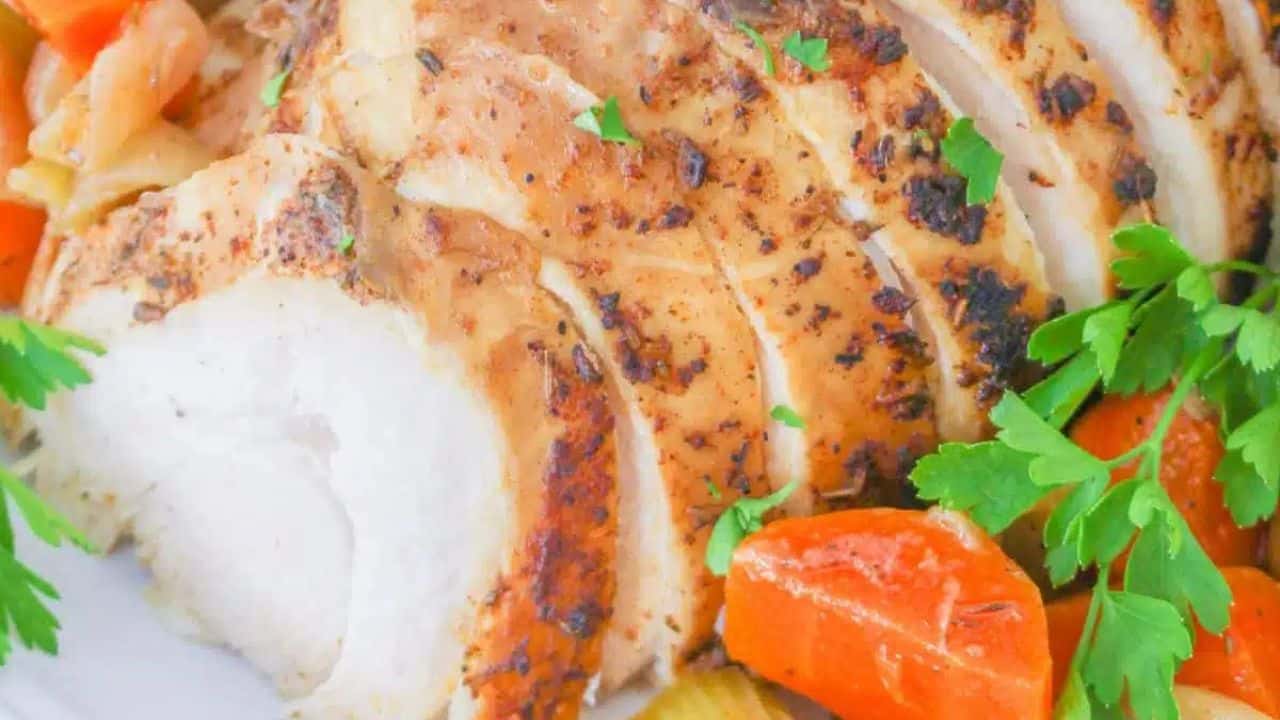 Sliced roasted chicken breast with herbs on an oval platter, surrounded by cooked carrots and celery. A small bowl of sauce is placed nearby.