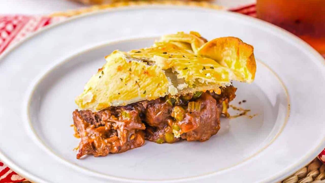 A close-up of a beef and vegetable pie with a golden, flaky crust. A triangular segment is removed, revealing chunks of beef and vegetables in a savory sauce inside the pie. The crust is lightly seasoned with herbs.