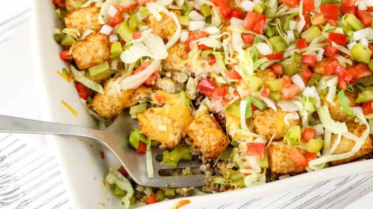 A casserole dish filled with tater tots, ground beef, diced tomatoes, shredded lettuce, onions, and pickles, with a serving spoon on the side.
