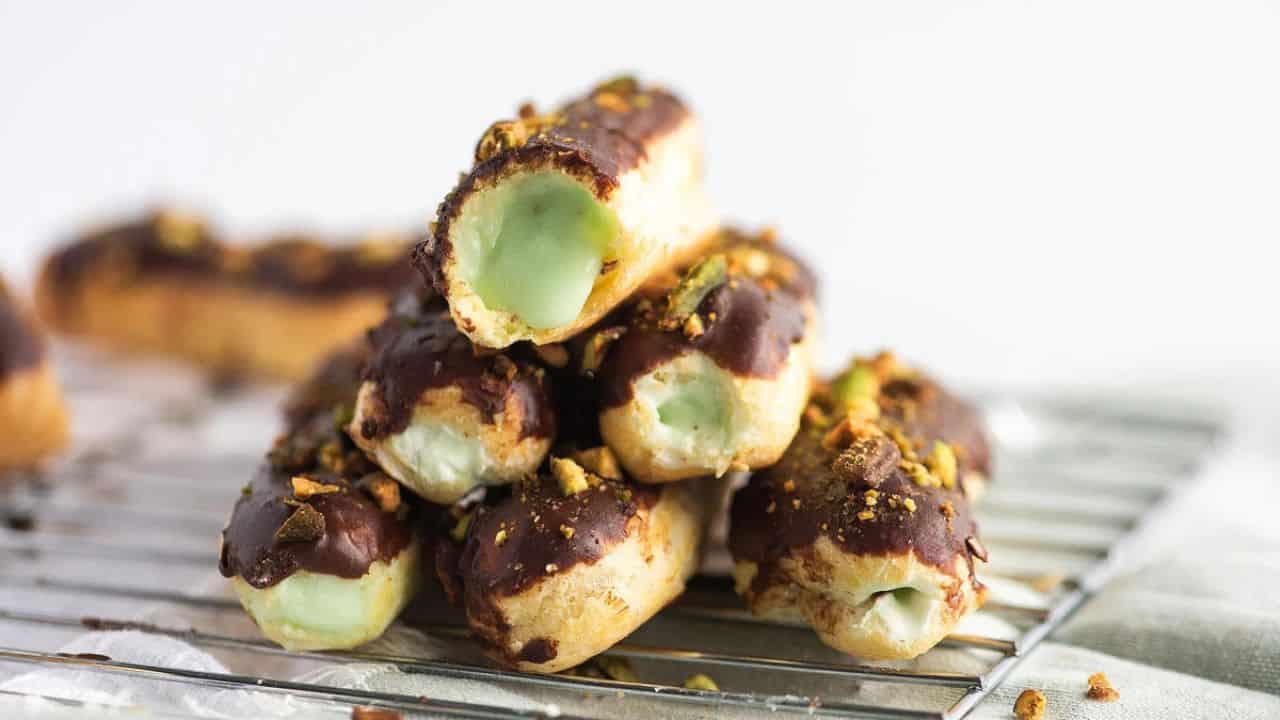 A pile of chocolate-covered éclairs filled with green pistachio cream rests on a wire cooling rack, with chopped pistachios sprinkled on top.