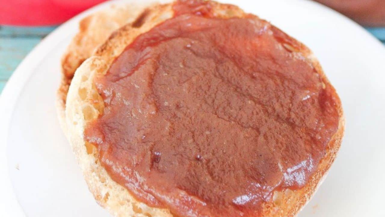A toasted English muffin half spread with a layer of apple butter sits on a white plate.